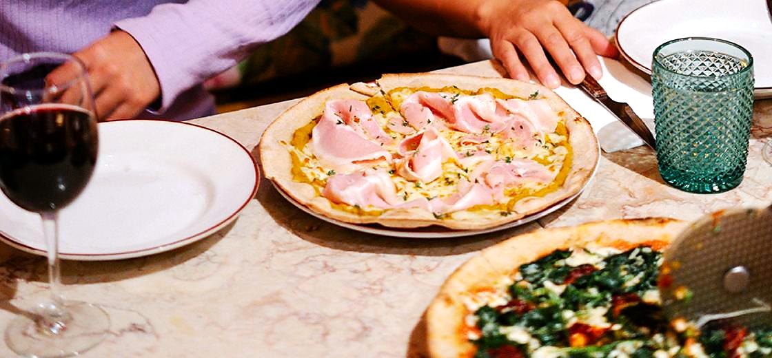 Mesa com pizzas acabadas de servir, um copo de vinho tinto e copos verdes lavrados