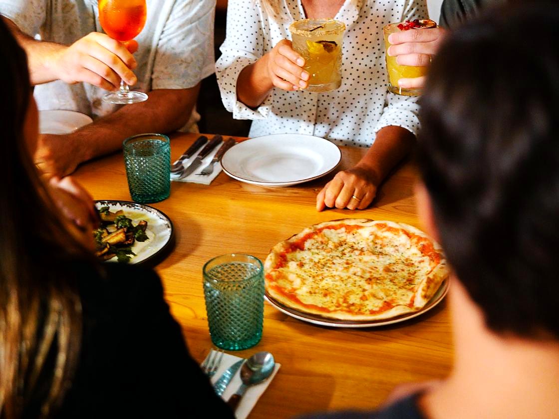 Mesa de grupo com cocktails erguidos num brinde, pizza e pratos para partilhar